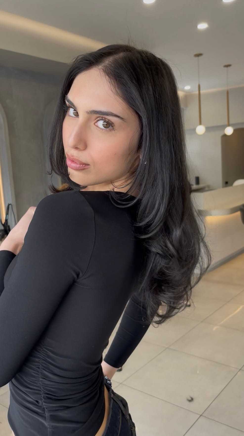 Woman with long dark hair wearing a black top, standing in a modern, stylish room.