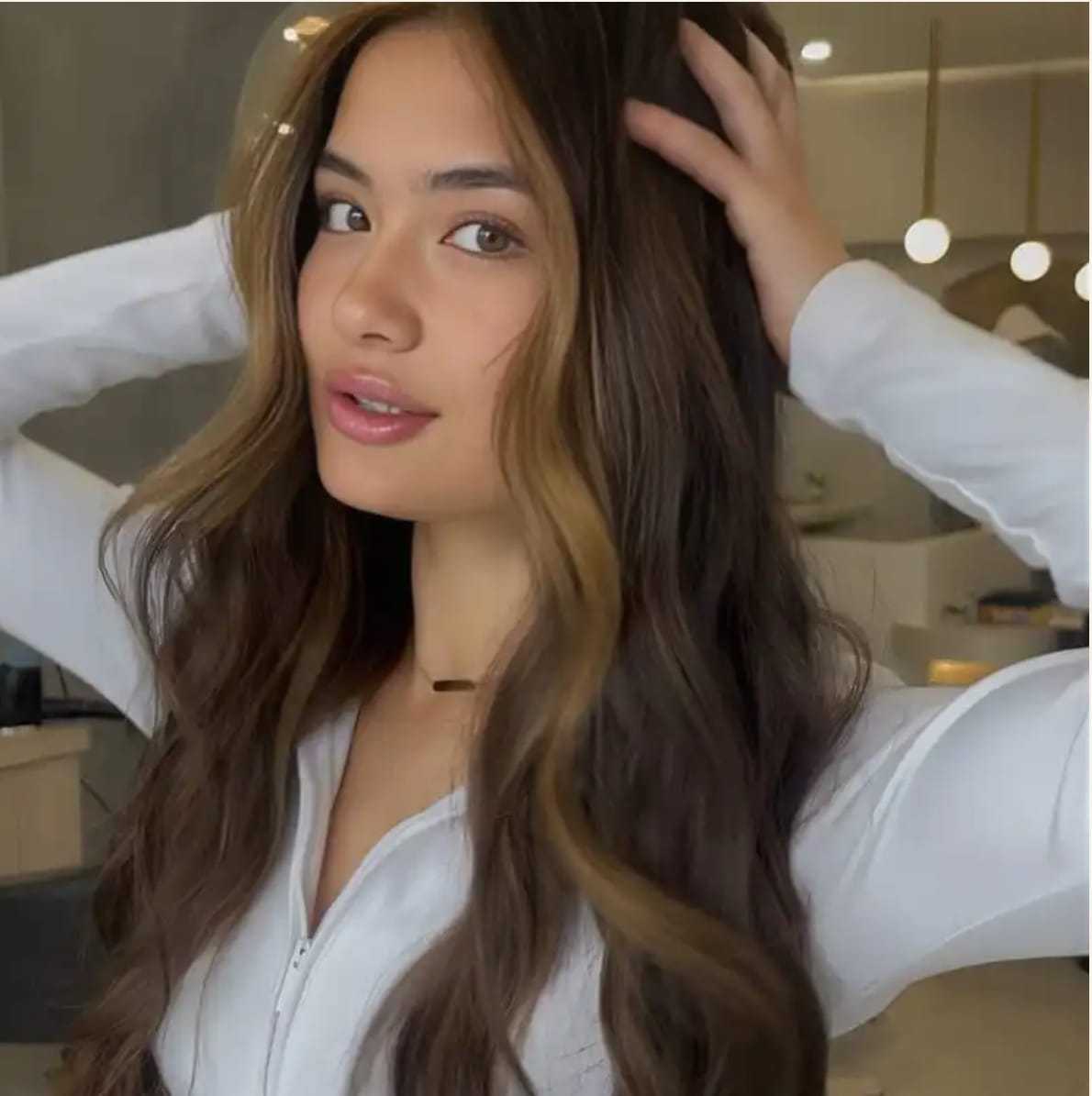 Young woman with long, wavy hair poses in a modern, well-lit room.
